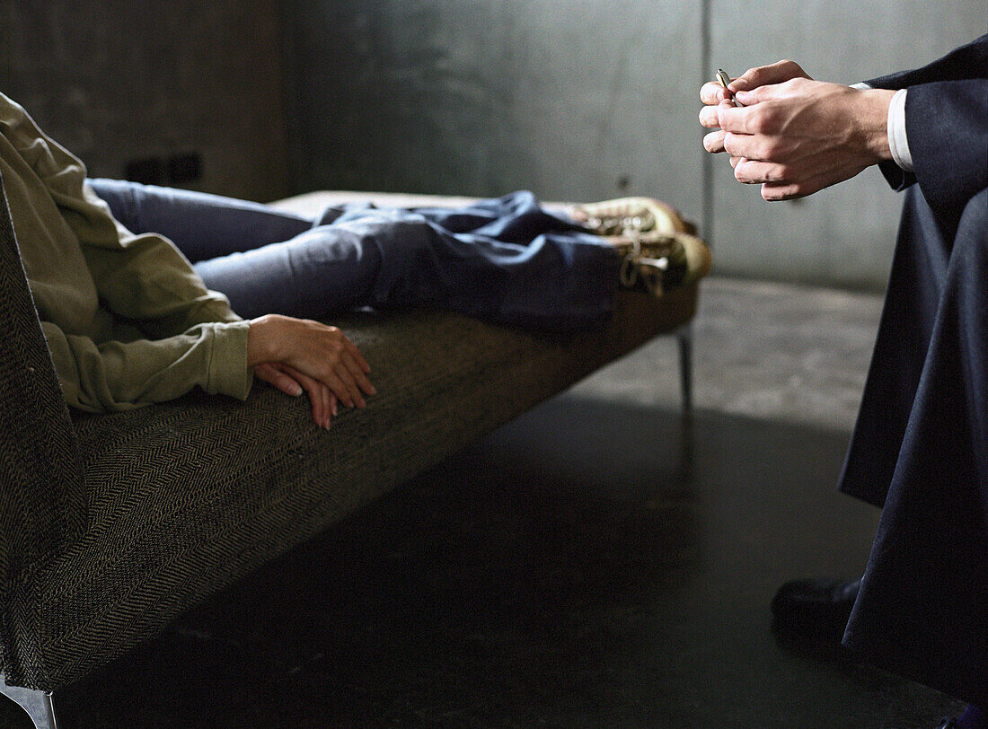 Young woman lying on couch, therapist leaning toward her with hands clasped, cropped