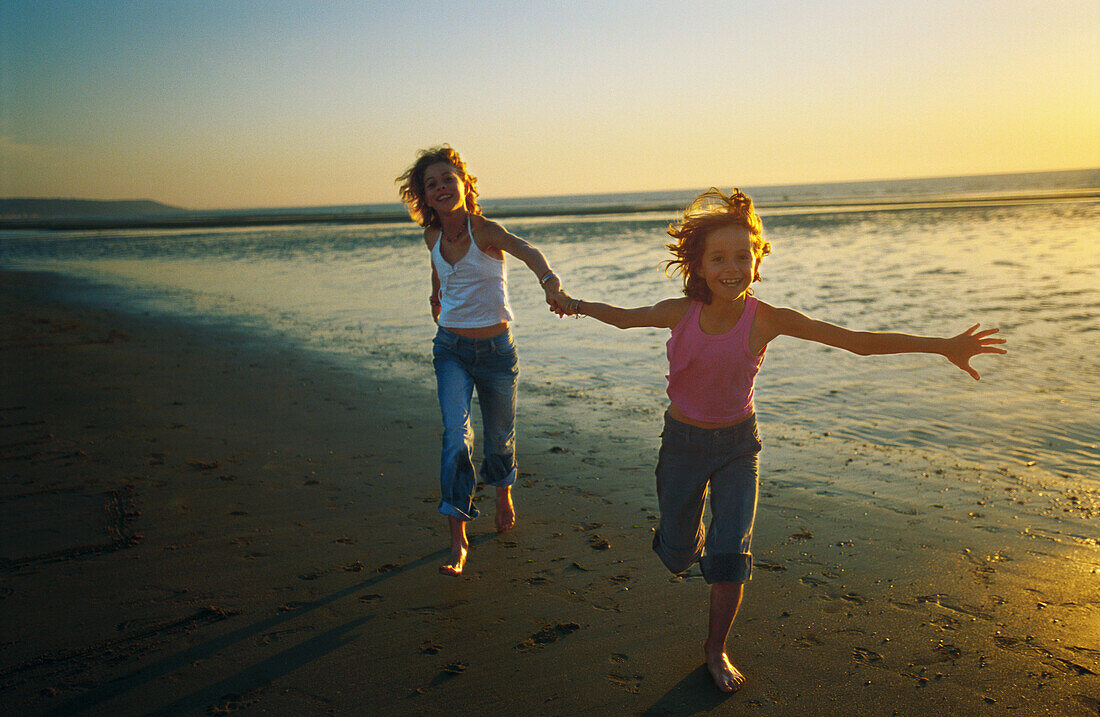 Two girls running hand in hand on beach, smiling