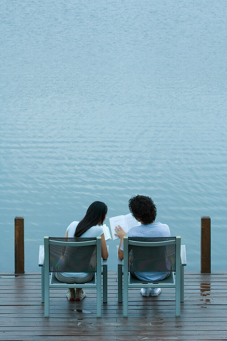 Couple sitting side by side on dock, reading books, rear view