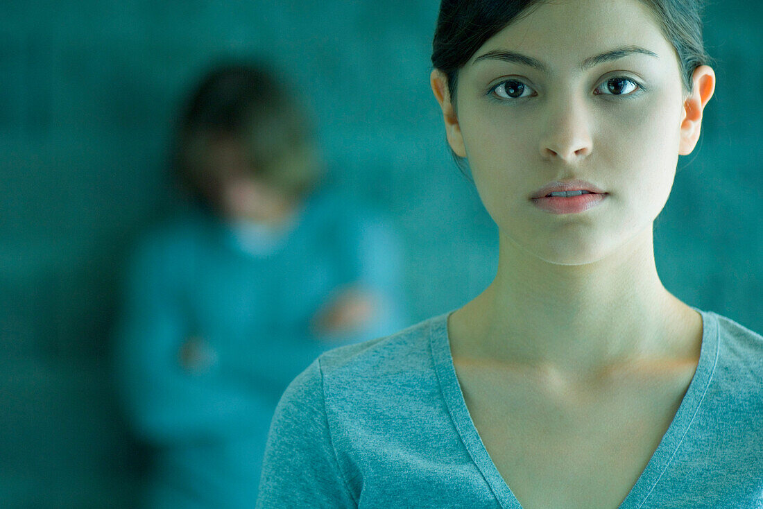 Young woman looking at camera, man in background leaning against wall with arms folded and head lowered