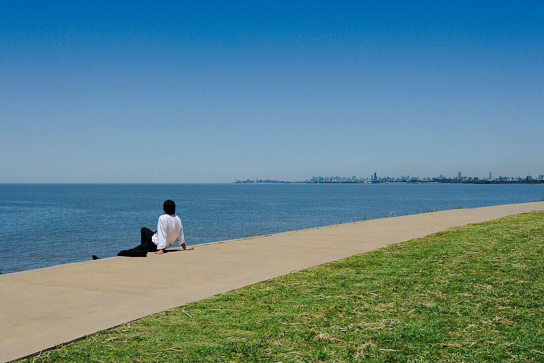 Man sitting on sidewalk beside sea, looking at horizon
