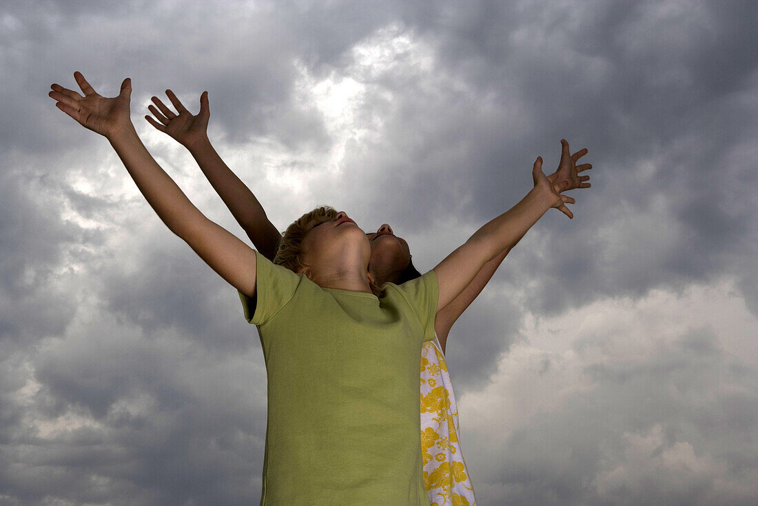Boy and girl with arms raised, heads back looking up at cloudy sky