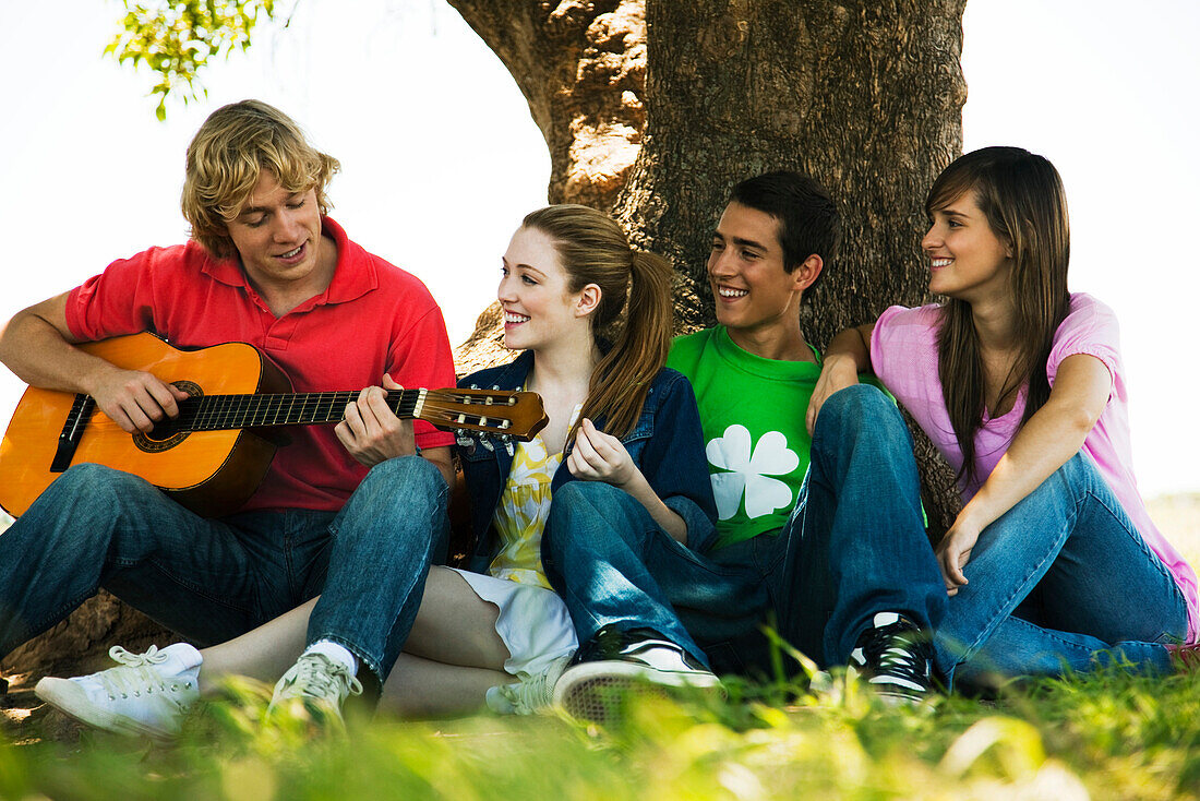 Young man sitting with friends under tree, playing guitar