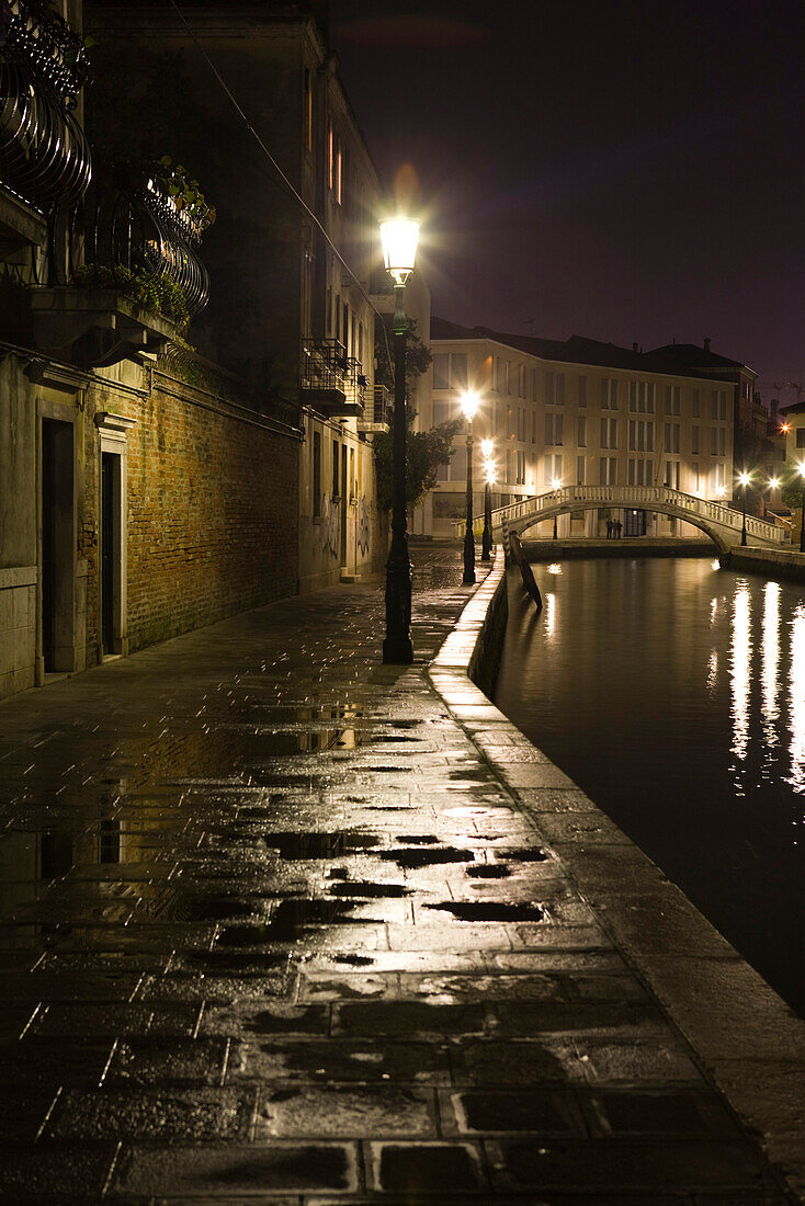 Italy, Venice, sidewalk along canal illuminated by street lamps at night