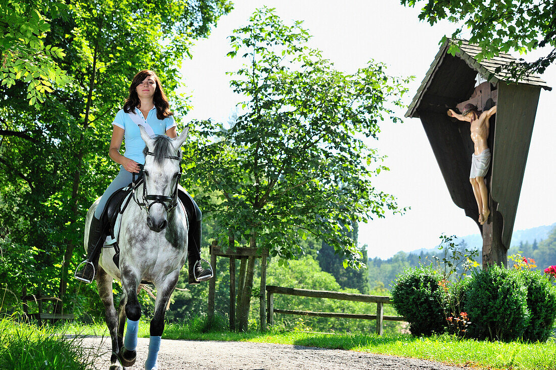 Woman riding a horse passing a wayside cross, Inn Valley, Upper Bavaria, Bavaria, Germany