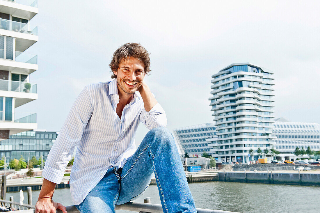 Mid adult man smiling at camera, Marco-Polo Tower in background, HafenCity, Hamburg, Germany