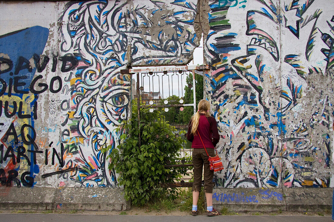 Germany, Berlin, Friedrichshain, East Side Gallery, teenager girl looking through the Wall, rear view