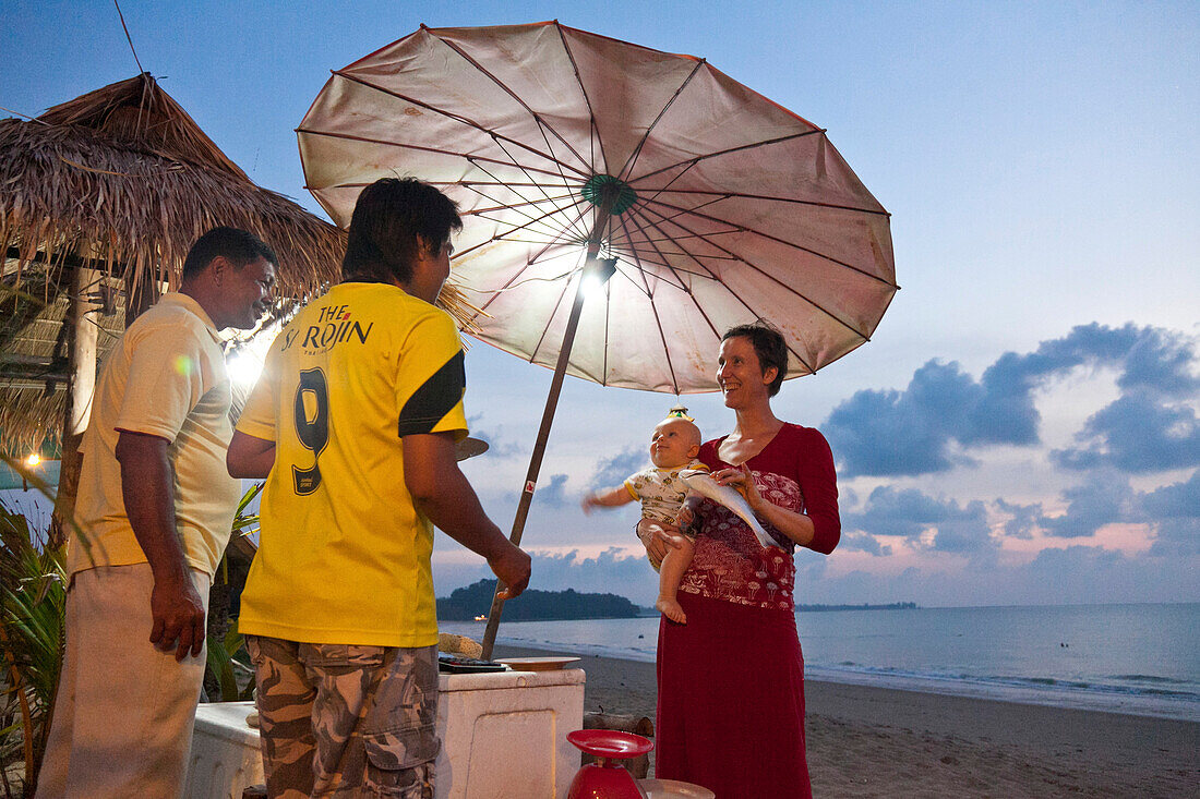 Deutsche Reisende mit Baby auf dem Arm kauft einen Fisch von zwei Thailändern, Strand in der Abenddämmerung, Pad Veek Beach, Andamanensee, Indischer Ozean, Bakseng Beach, Khao Lak, Thailand, Asien