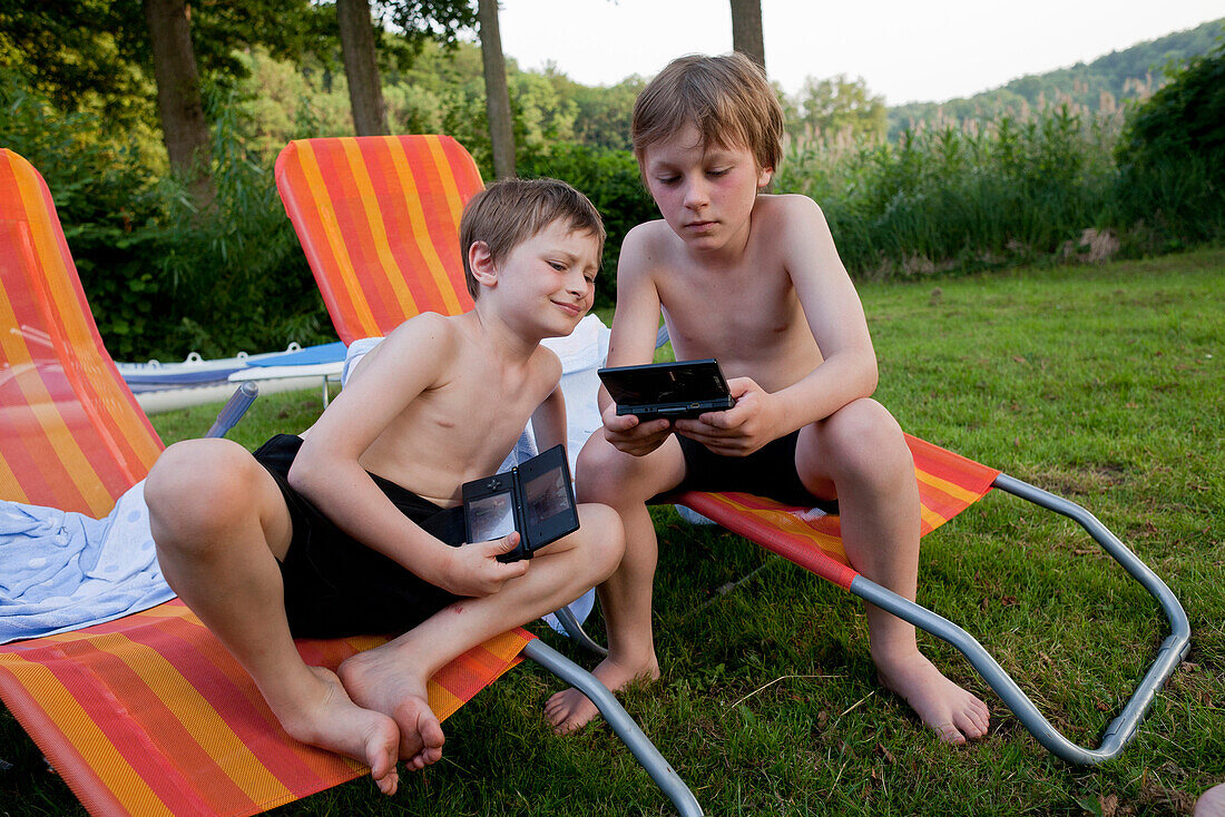 Two boys sitting on deckchairs playing with handheld video game, Lankau, Schleswig-Holstein, Germany