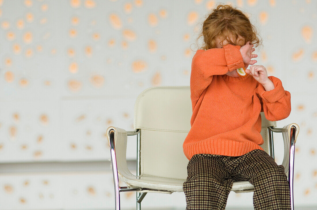 Little girl sitting on a chair, covering her face with her arm