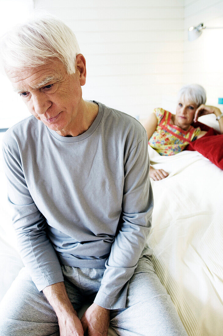 Sad looking senior man sitting on a bed, senior woman in background