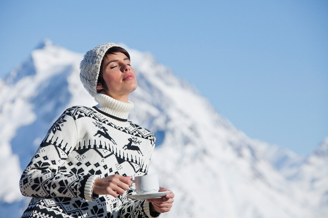 Young woman with coffee cup, enjoying winter sun, mountains in background