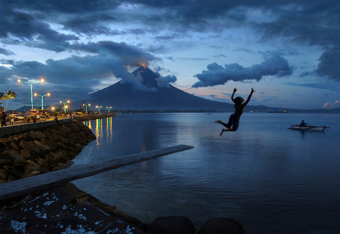 Boy jumping into the sea before a glowing Mayon Volcano, Legazpi City, Luzon Island, Philippines, Asia
