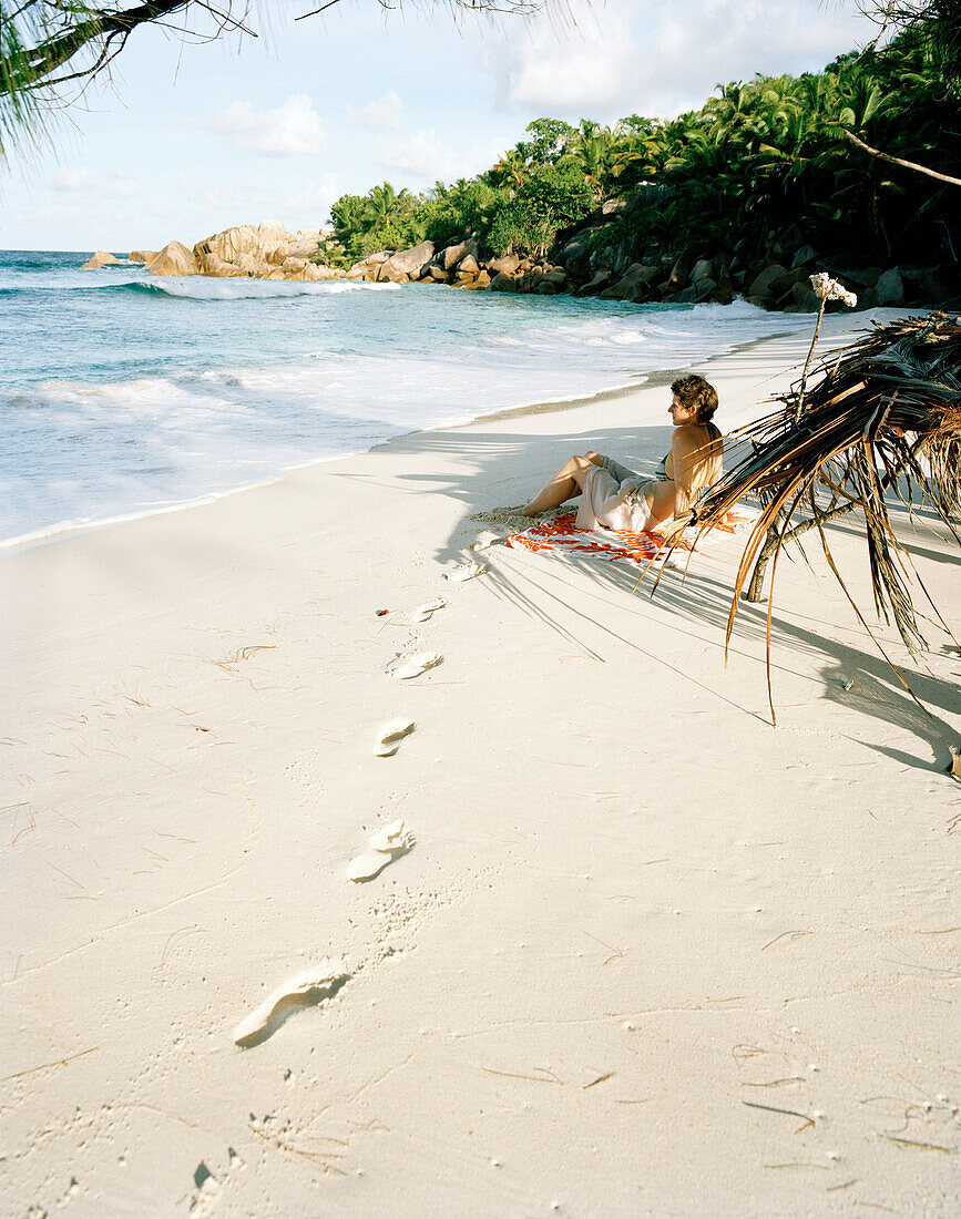 Frau sitzt allein am Strand Anse Cocos, im Süden von La Digue, La Digue and Inner Islands, Republik Seychellen, Indischer Ozean