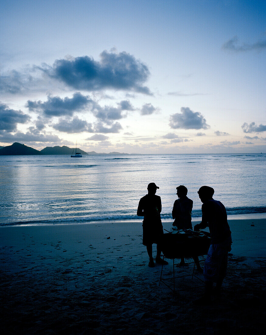 Fischgrillen auf kreolische Art mit Öltonnengrill am Strand am Abend, La Passe, im Hintergrund Nachbarinsel Praslin,  La Digue, La Digue and Inner Islands, Republik Seychellen, Indischer Ozean