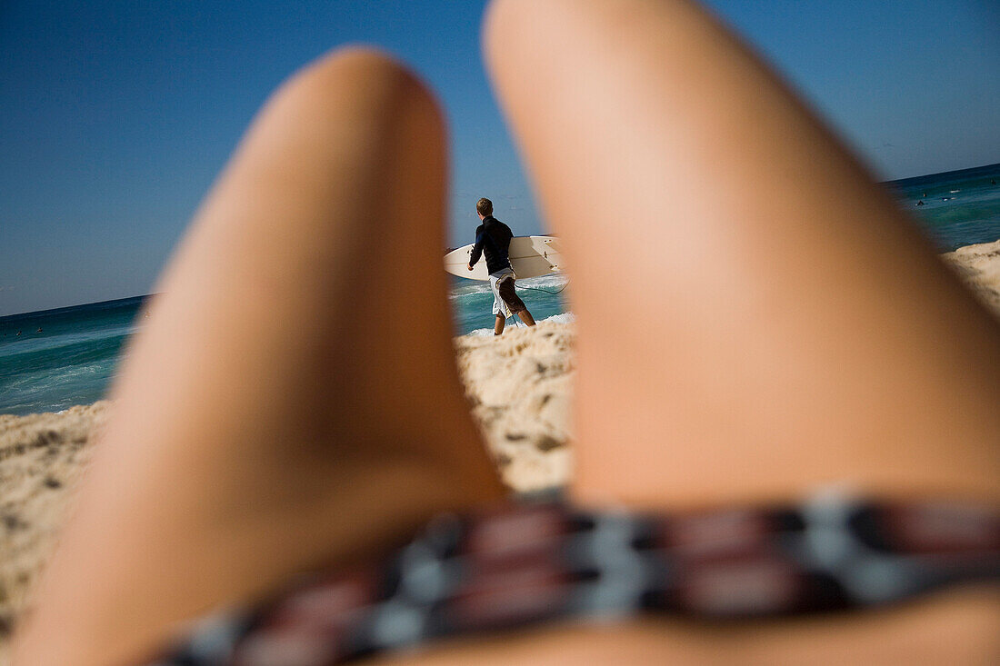 Surfer viewed through womans legs lying on Bondi Beach, Sydney, New South Wales, Australia