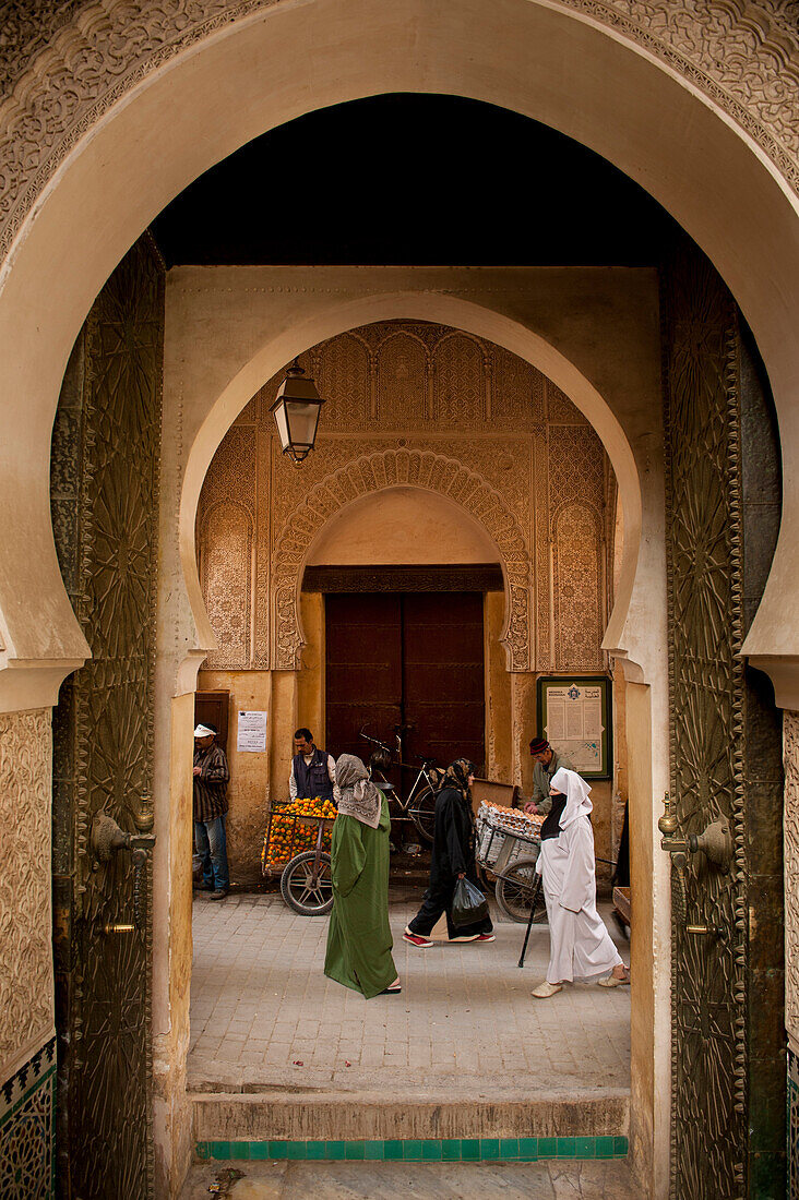 People walking past orange and egg sellers outside entrance to Medersa Bou Inania, Fez, Morocco