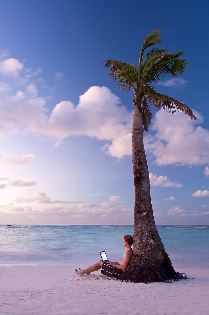 Man using laptop beside palm tree at dawn on Worthing Beach, Barbados, Barbados