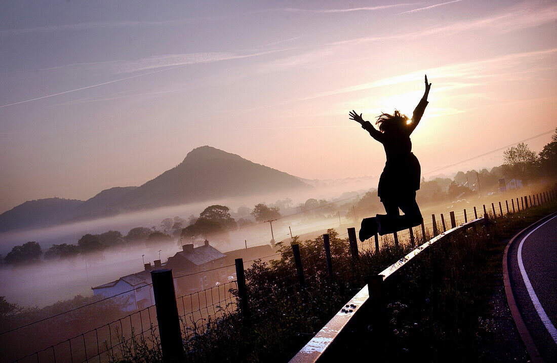 Girl jumping on a country road at dawn, Powys, Wales