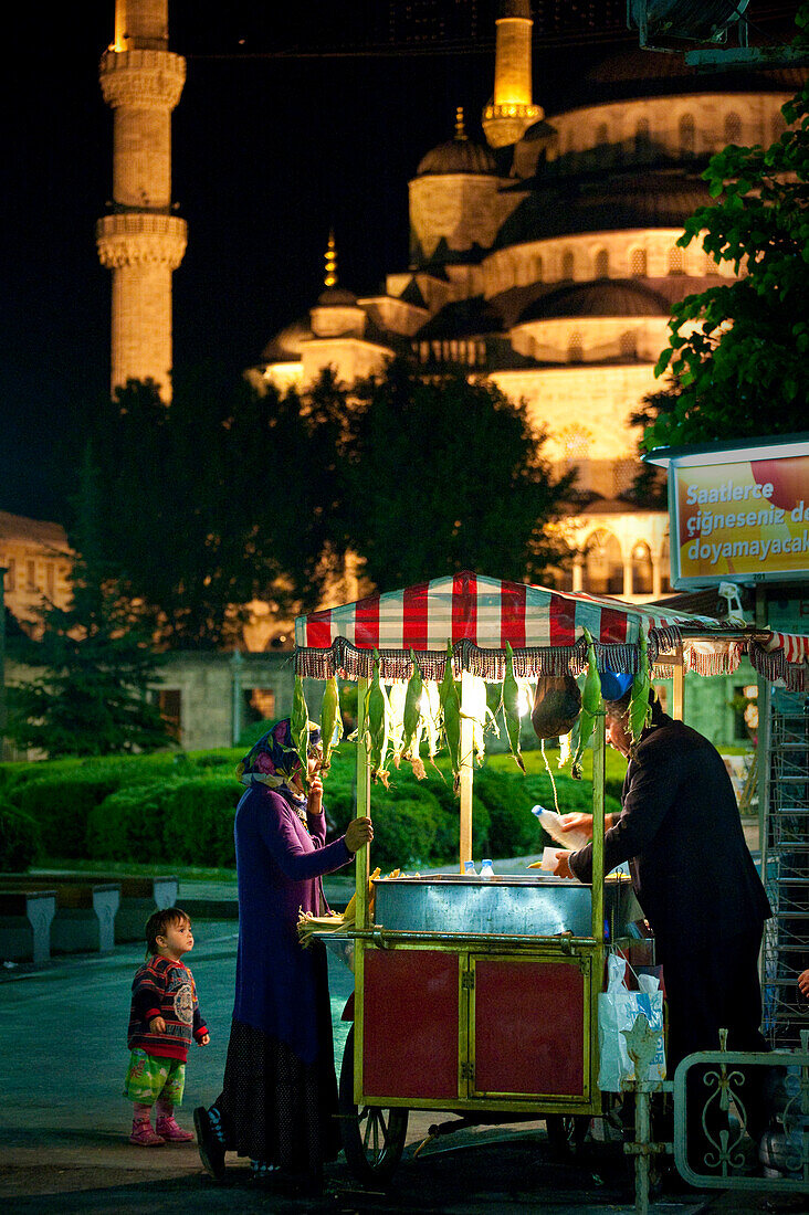 Mother and child buying from sweetcorn seller in front of the Sultanahmet or Blue mosque at night, Istanbul Turkey