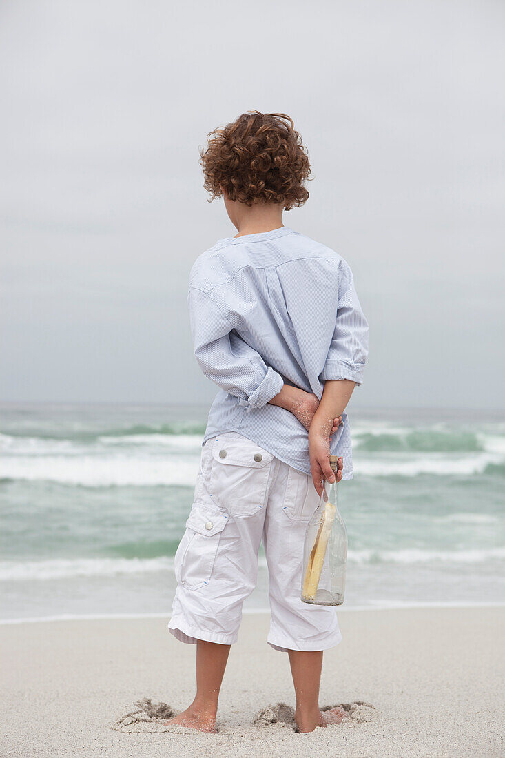 Rear view of a boy holding bottle with message on the beach