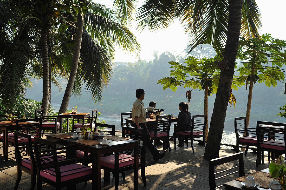 Restaurant terrace under palm trees at Nam Khan river, tributary to Mekong river, Luang Prabang, Laos, Lao Peoples Democratic Republic