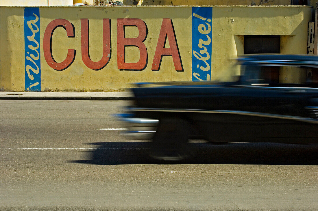 A blurred classic car passes a Viva Cuba sign, Havana, Cuba.