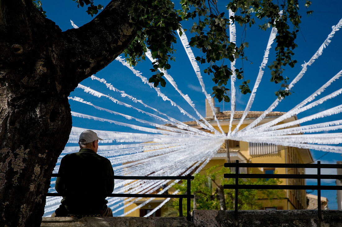 Man sitting by tree in front of white bunting in Caimari square, rear view, Majorca, Ballearic Islands, Spain
