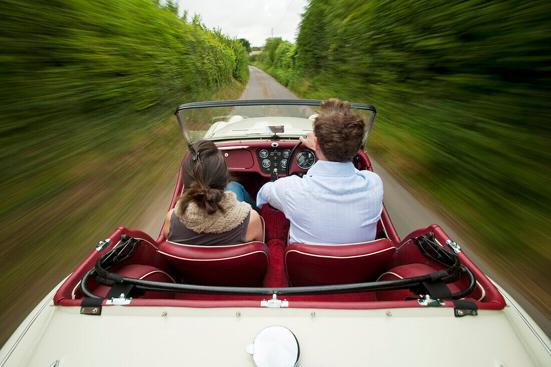 Couple driving a convertible car on a hedge lined country road / lane, East Sussex, England