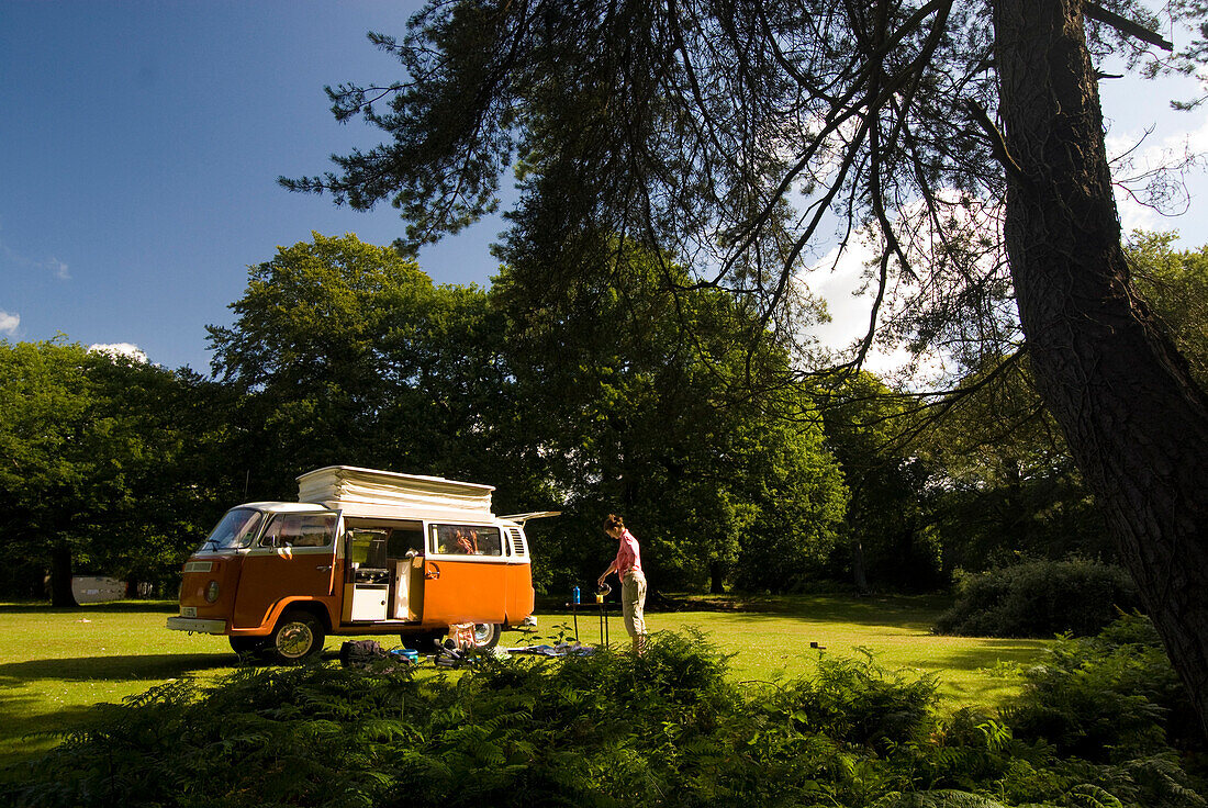 Woman making tea outside old campervan in forest clearing, New Forest, Hampshire, UK