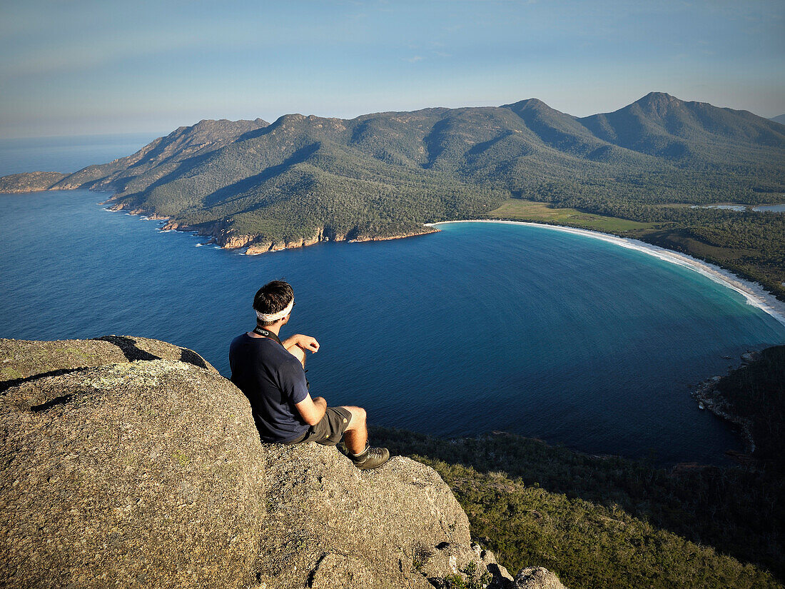 Wanderer genießt Blick von Fels des Mount Amos auf Wineglass Bay, Freycinet Nationalpark, Tasmanien, Australien