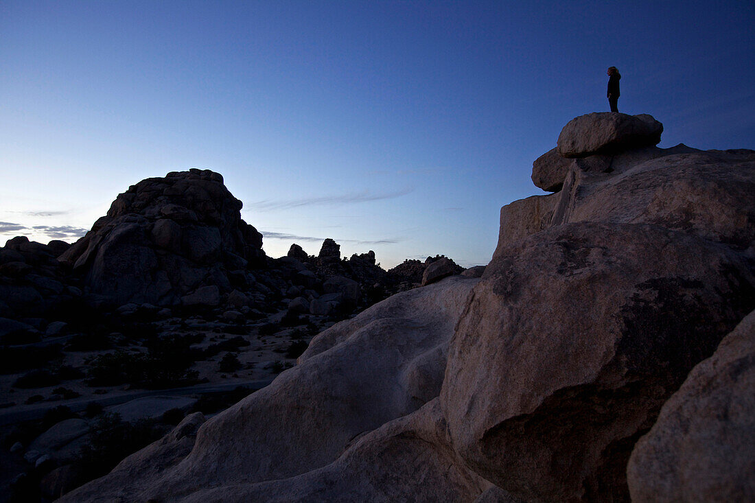 Junge Frau steht auf einem Felsen im Joshua Tree National Park und genießt die Abenddämmerung, Joshua Tree National Park, Kalifornien, USA