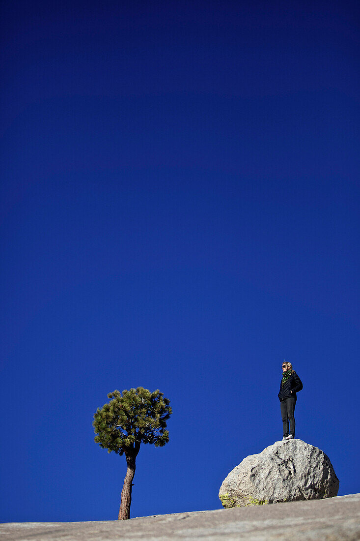 Junge Frau steht auf einem kleinen Felsen neben einem Baum, Yosemite-Nationalpark, Kalifornien, USA