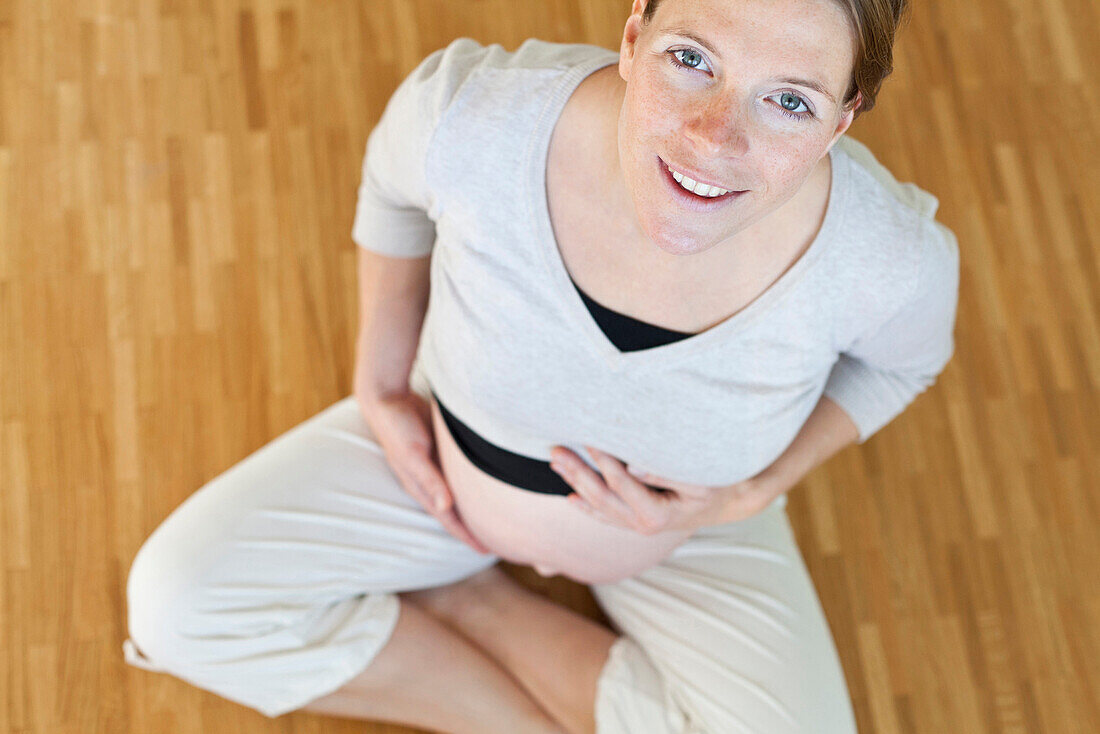 Schwangere junge Frau hält dicken Bauch, sitzt auf Parkett im Wohnzimmer, schwanger im 8. Monat, Blick in die Kamera, MR, Leipzig, Sachsen, Deutschland