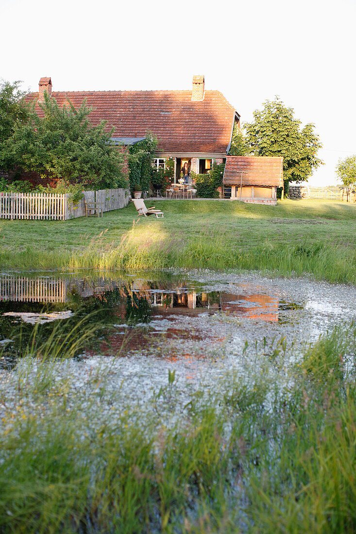 Blick über einen Teich auf eine Haus mit Garten am Abend, Haus Strauss, Bauernkate in Klein Thurow, Roggendorf, Mecklenburg-Vorpommern, Deutschland