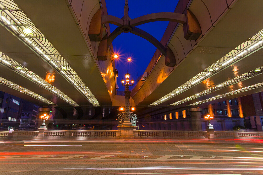 A slow shutter speed captures the light streaks of traffic crossing venerable Nihonbashi Bridge with elevated expressways overhead at twilight in the Nihonbashi District of central Tokyo, Japan.
