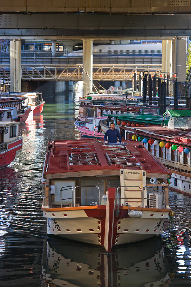 With a Shinkansen bullet train rushing past in the background, a boat captain eases his yakatabune excursion houseboat past others moored in a narrow canal just off the Sumida River in the Hamamatsucho District of Tokyo, Japan.
