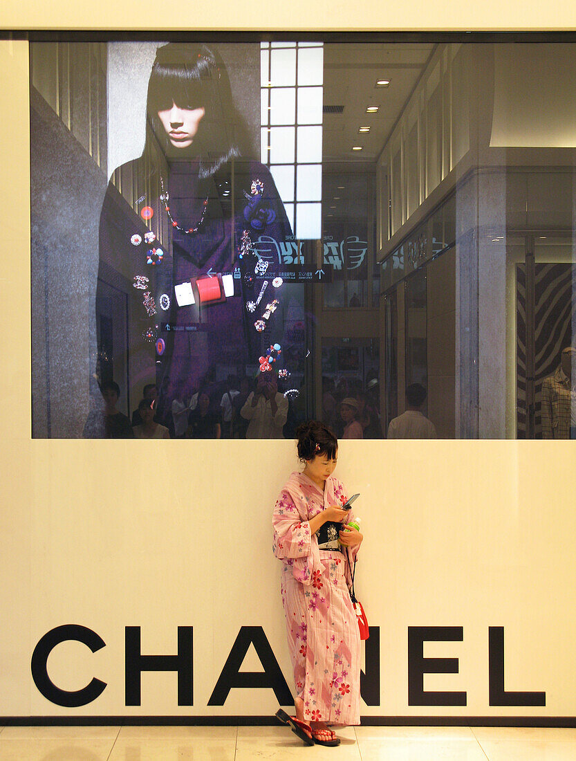 A young Japanese woman wearing a yukata traditional summer kimono uses her cellphone while casually leaning against a huge wall-size Chanel advertisement at Takashimaya Times Square department store in the Shinjuku district of Tokyo, Japan.