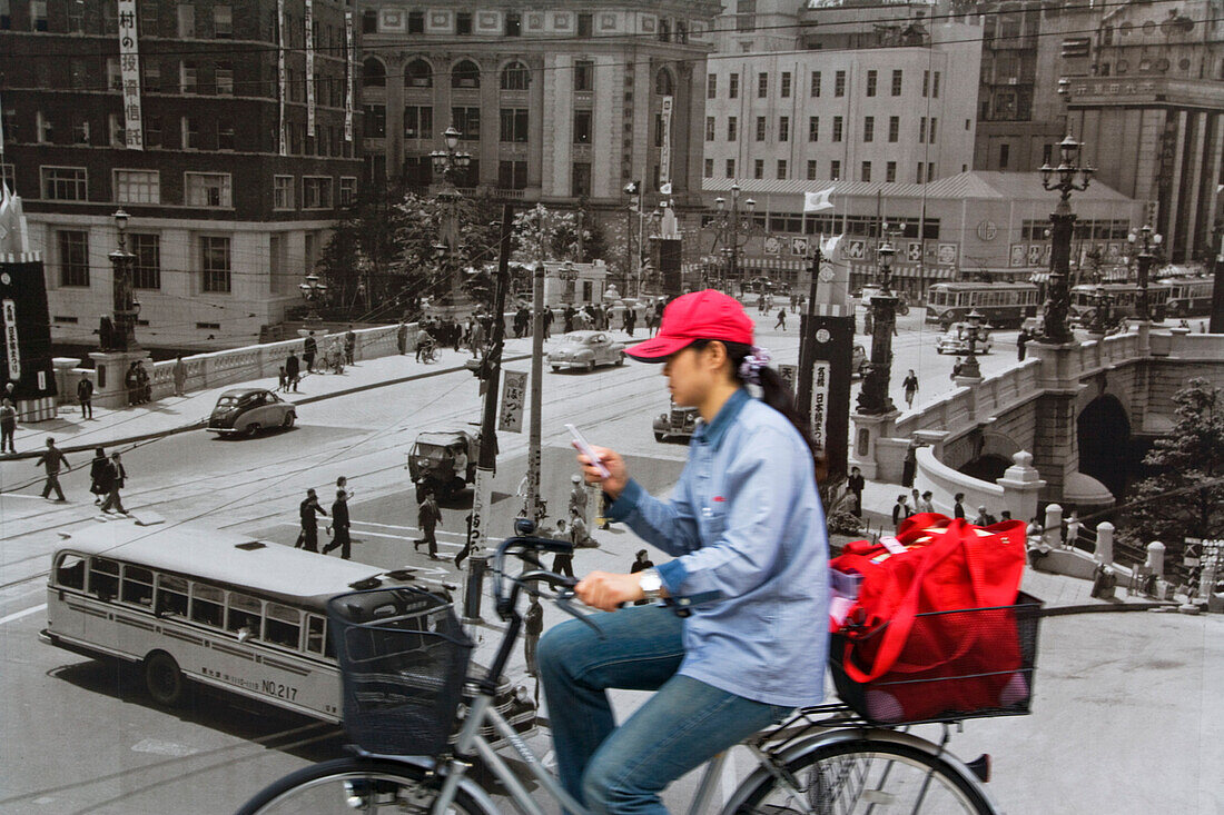 A young Japanese woman uses her cellphone while riding a bicycle past an old 1950's black-and-white mural of Nihonbashi Bridge at a construction site in the Nihonbashi District of central Tokyo, Japan.