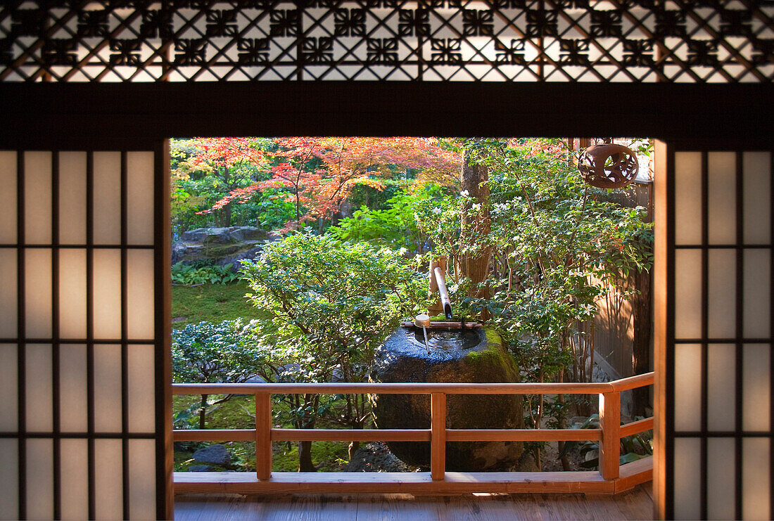 A detailed view looks out through washi-paper shoji doors beneath a decorative transom to the stone tsukubai water basin by the garden at Daiho-in, a sub-temple inside Myoshinji Temple complex, located in Kyoto, Japan.