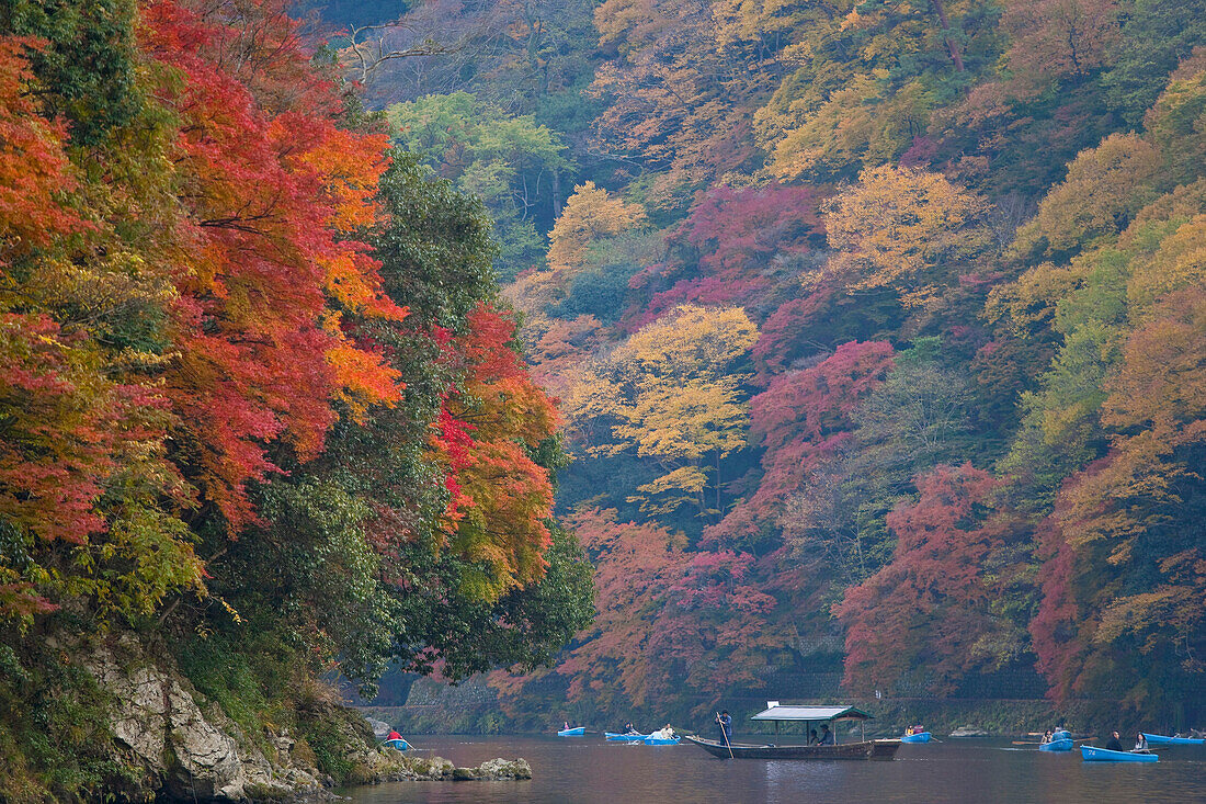 A small excursion boat floats down the Hozu River valley amidst rental rowboats and a palette of autumn hues under diffuse light in the northwest Arashiyama district of Kyoto, Japan.