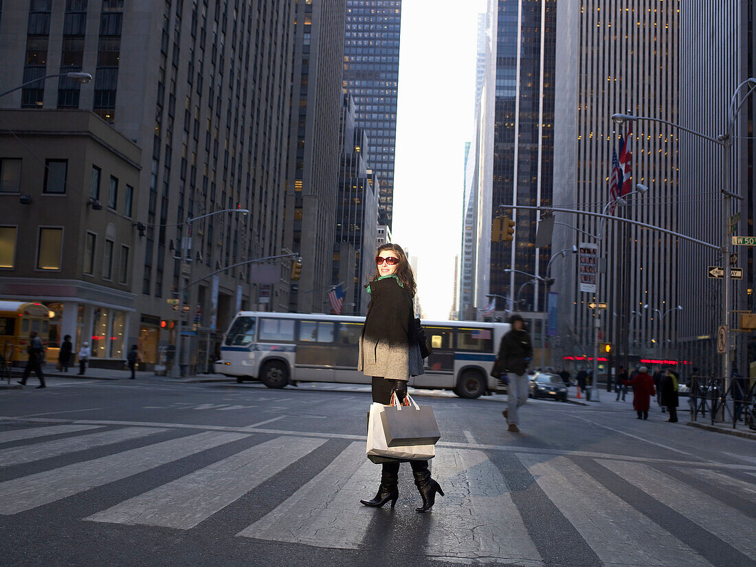 Elegant woman crossing New York street. Woman with shopping bags crossing New York street. Sky scrapers behind her. She is looking at camera, smiling.