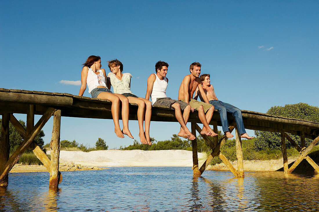 Group of young people sitting on jetty. Two teenage girls and three young men are relaxing in the sun on jetty, they are smiling.