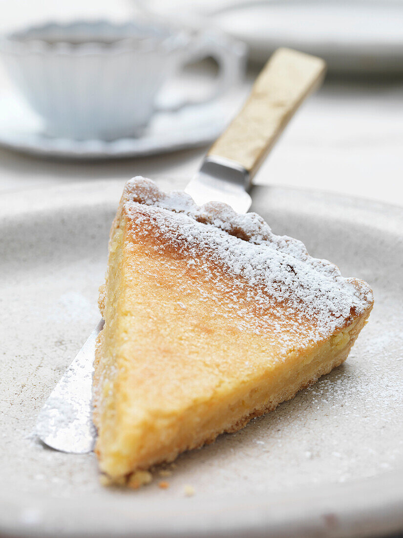 Slice of lemon tart on plate. Lemon Tart dusted with icing sugar neutral plate with cup of coffee in the background