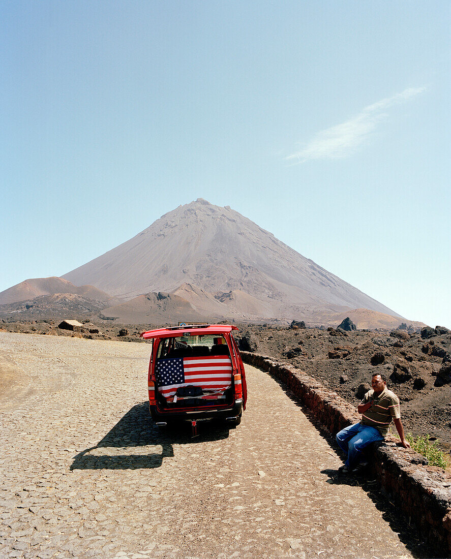 Fahrer und Kleinbus mit USA Flagge auf der Strasse vor dem Vulkan Pico de Fogo, 2.829 m hoch, Cha das Caldeiras, Insel Fogo, Ilhas do Sotavento, Republic Kap Verde, Afrika