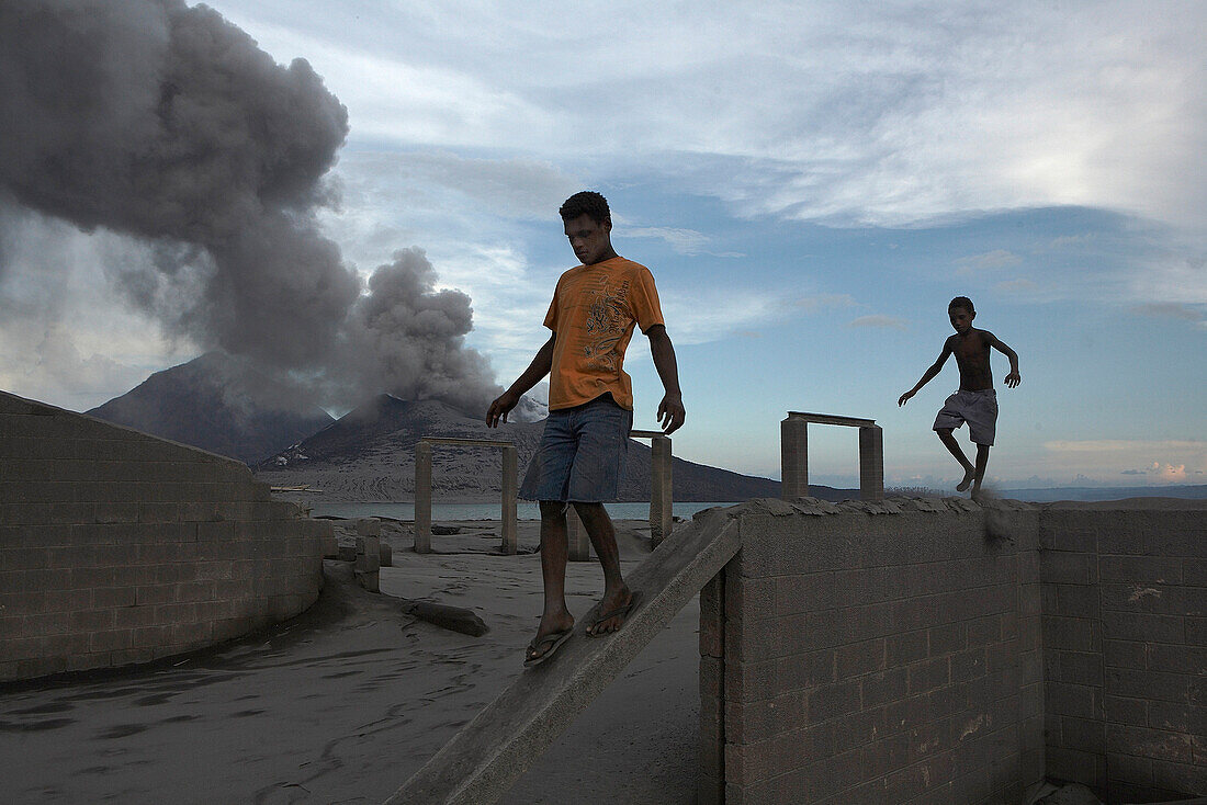 Children playing in the ruins of an old cement factory with erupting Volcano in the background. Tavurvur Volcano, Rabaul, East New Britain, Papua New Guinea, Pacific