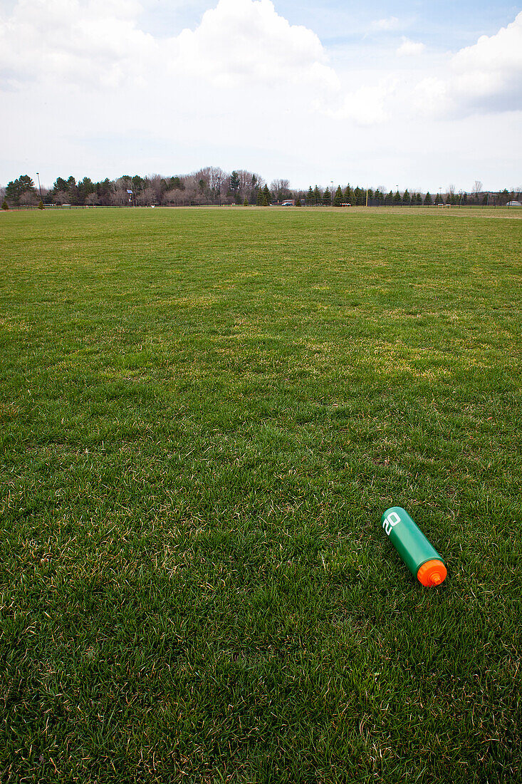 A sports ground, grass field, with trees on the horizon.  A water bottle on the ground., A Water Bottle on a Sports Field