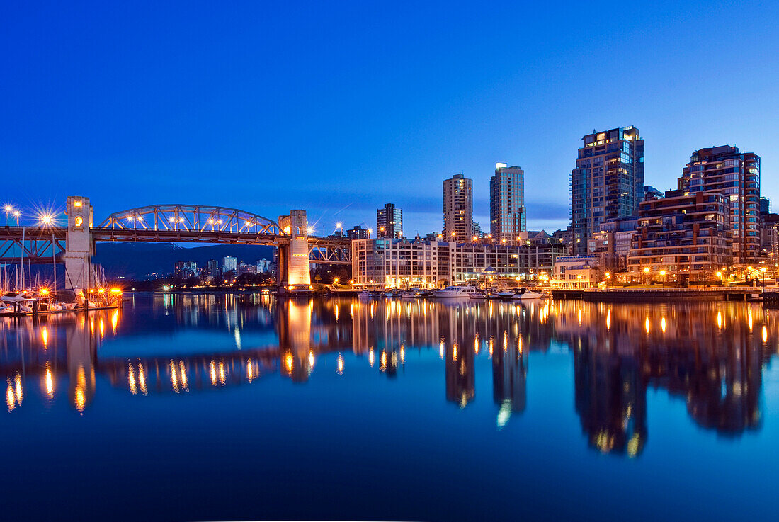 Skyline Reflected in False Creek. The Vancouver skyline at twilight or dawn, with low light and blue sky, and flat calm water. Lit up skyline and tall buildings. British Columbia, Canada., Vancouver Skyline Reflected in False Creek