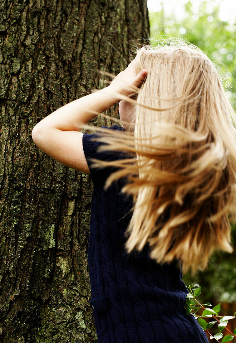 Young Blond Girl Standing Against Tree Trunk, Rear View