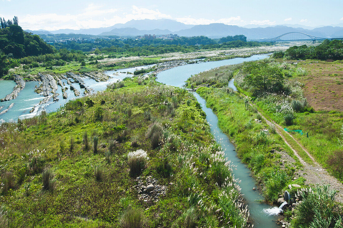 Irrigation channels and a drainage overflow ditch cut into the landscape by the river. Agricultural land and water management. Overflow pipes. Landscape. Bridge and mountains in the distance., Taoyuan, Taiwan, Asia.